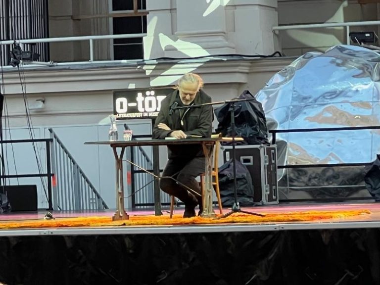 Mann in schwarzer Kleidung liest an einem Tisch auf der ImPulsTanz Bühne in Wien, sichtbar ist die Bühnen- und Tontechnik von Eventpower.