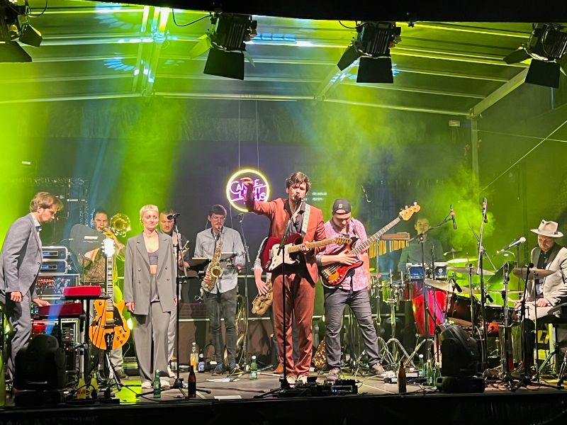 Band mit Gitarre, Schlagzeug, Bläsern und Sängern auf Bühne mit sichtbarer Tontechnik, Licht- und Bühnentechnik.